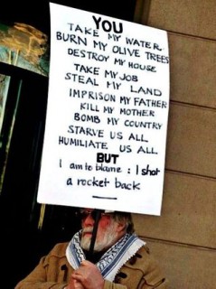 An Elderly Palestinian Man Carries a Placard of the Truth.  It says: “You take my water, burn my olive trees, destroy my house, take my job, steal my land, imprison my father, kill my mother, bombard my country, starve us all, humiliate us all but I am to blame: I shot a rocket back.”  